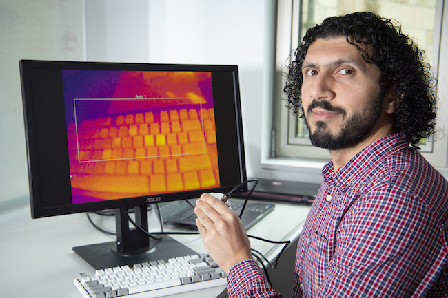Docent Mohamed Khamis z Fakulty výpočetní techniky Glasgowské university předvádí, jak malou termovizní kamerou v jeho levé ruce nasnímá teplotní obraz klávesnice, na které před chvílí psal. Tlačítka prozrazují nedávné doteky jeho prstů. Kredit: Univ