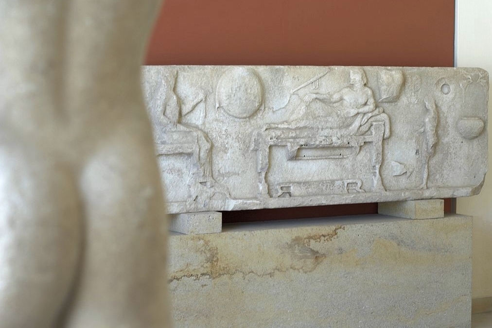 Archilochův reliéf na pozadí kúra A 1281. Úhel pohledu byl zvolen také jako ilustrace k Archilochově parodii na spartskou bojovou píseň. Archeologické muzeum na Paru, A 758. Kredit: Zde, Wikimedia Commons. Licence CC 3.0.