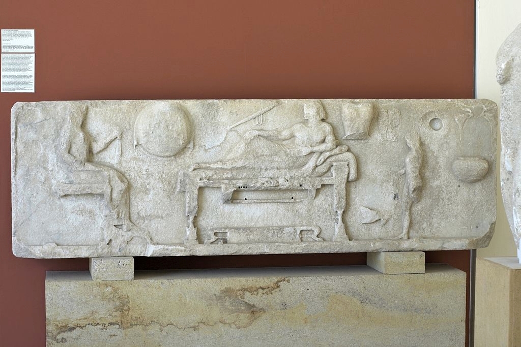 Reliéfní stéla z Archilocheia na Paru, kolem roku 500 před n. l. Archilochos leží na lehátku, vlevo sedí jeho žena, zprava obsluhuje chlapec. Na zdi visí štít, toulec a lyra. Archeologické muzeum na Paru, A 758. Kredit: Zde, Wikimedia Commons. Licenc