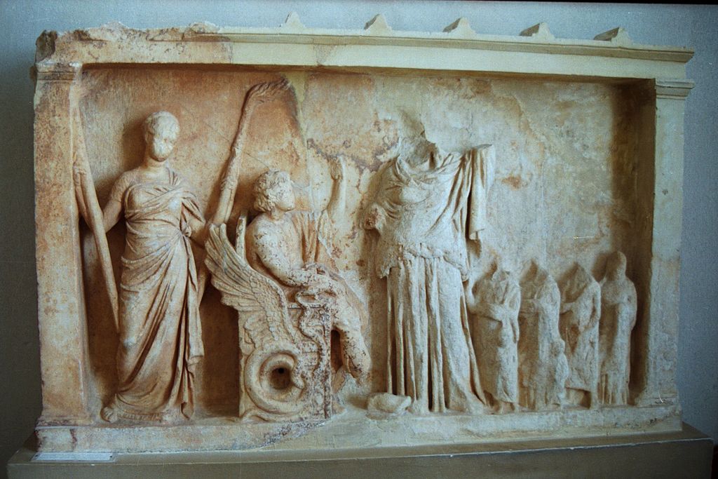 Triptolemos na dračím voze, asi se vypravuje distribuovat po světě setbu obilí, kterou dostal od bohyně. Archeologické muzeum v Eleusině. Kredit: Zde, Wikimedia Commons.