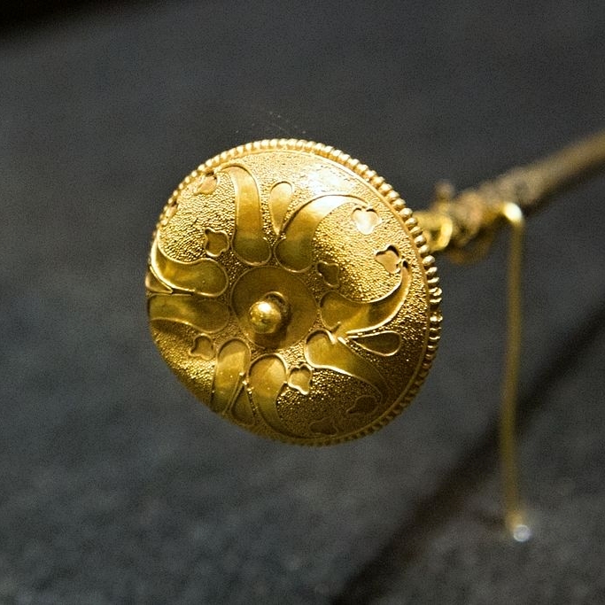 Ozdobný knoflík, smontován s novověkou jehlou do podoby jehlice. Horní plocha zdobená jemnou granulací, hladký podkladový plech, lemovaný filigránovými drátky, které vytvářejí rostlinný motiv. Zlato. Etrurie, konec 6. až 5. století před n. l. NM-H10 