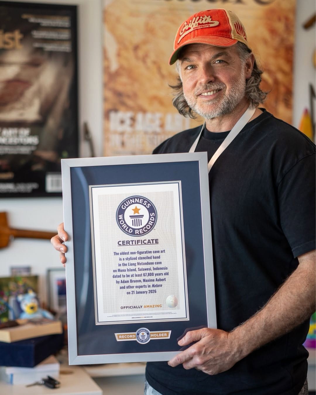 Sympaťák Adam Brumm.  Na snímku s certifikátem od Guinness dokládajícím „Nejstarší malbu nefigurativního umění“.  Tento jeho objev se týká obtisku ruky který v indonéské jeskyni zanechal člověk před nejméně 67 800 lety. (Kredit: Griffith University).