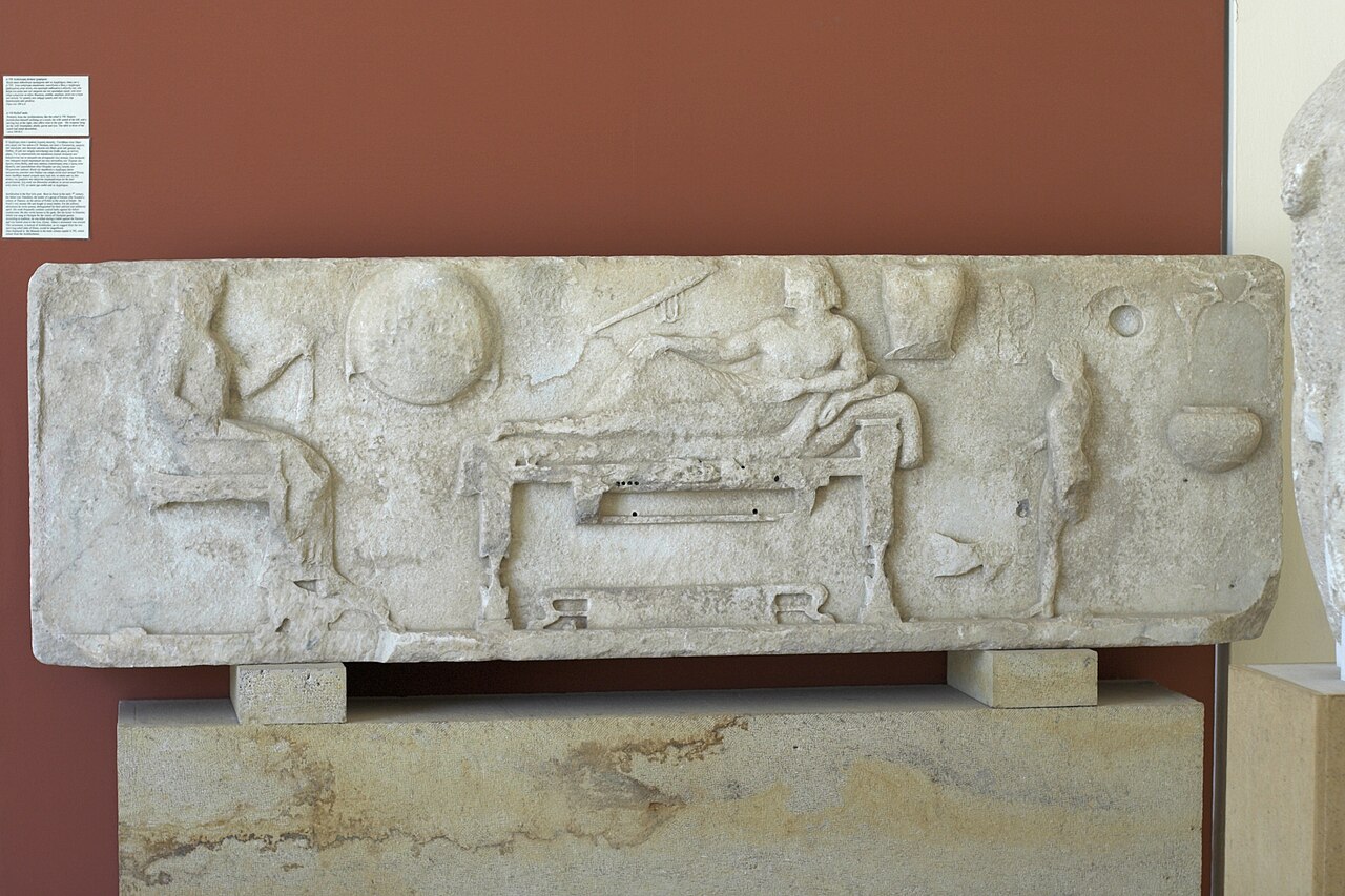 Reliéfní stéla z Archilocheia na Paru, kolem r. 500 před n. l. Archilochos leží na lehátku, vlevo sedí jeho žena, zprava obsluhuje chlapec. Na zdi visí štít, toulec a lyra. Archeologické muzeum na Paru, A 758. Kredit: Zde, Wikimedia Commons. Licence 