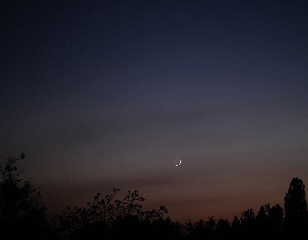 Zapadající srpek mladého Měsíce (je mu 43 hodin po novu) s Merkurem (vpravo) při začátku nautického soumraku, asi 7 stupňů nad obzorem, v květnu 2022. Disk Měsíce je doplněn popelavým svitem, tedy světlem Země. Vlevo dole je hvězdný obr Aldebaran. Kr
