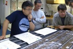Yuki Morono (vlevo) a Steven D'Hondt (zcela vpravo) na palubě výzkumného vrtného zařízení JOIDES Resolution. Před nimi jsou  jádra sedimentů získaná v jižním Pacifiku. Kredit: IODP JRSO.