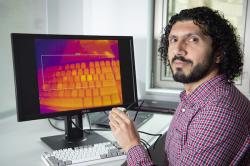 Docent Mohamed Khamis z Fakulty výpočetní techniky Glasgowské university předvádí, jak malou termovizní kamerou v jeho levé ruce nasnímá teplotní obraz klávesnice, na které před chvílí psal. Tlačítka prozrazují nedávné doteky jeho prstů. Kredit: University of Glasgow