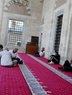 Muži si před hlavním vstupem do mešity mají možnost si odpočinout na kobercích. Někteří při tom mobilují, mnozí si tam přes poledne zdřímnou (fotit spící a vleže, nám bylo stydno). Kredit: Osel