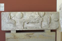 Reliéfní stéla z Archilocheia na Paru, kolem r. 500 před n. l. Archilochos leží na lehátku, vlevo sedí jeho žena, zprava obsluhuje chlapec. Na zdi visí štít, toulec a lyra. Archeologické muzeum na Paru, A 758. Kredit: Zde, Wikimedia Commons. Licence CC 4.0.